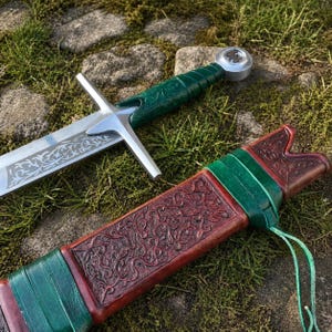 May include: A medieval-style sword with a silver blade and a green handle. The blade has an etched design. The sword rests on a bed of grass next to a brown leather sheath with green accents and an embossed pattern.