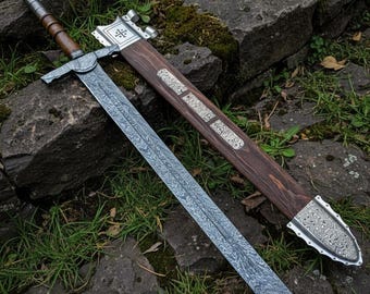 May include: A large, ornate sword with a patterned blade and a wooden handle and sheath. The sword's blade is silver with a detailed design. The sheath is brown with silver accents. The sword is lying on a stone surface.