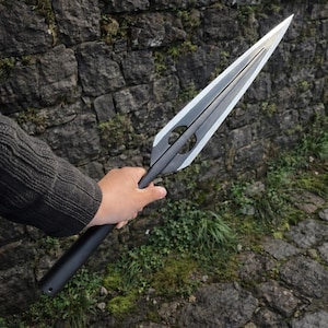 Puede incluir: Una lanza negra y plateada con una hoja larga y puntiaguda. La lanza tiene un mango oscuro y un diseño simétrico con dos recortes circulares cerca de la hoja. La lanza se sostiene contra una pared de piedra.