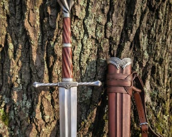 May include: A steel sword with a polished blade, a twisted brown wooden grip, and silver-toned metal accents on the crossguard and pommel. Beside the sword is a brown leather scabbard featuring silver-toned metal hardware and leather straps.