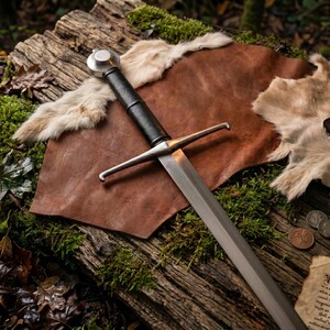 May include: A longsword with a polished steel blade, a straight crossguard, and a black leather-wrapped grip. The sword features a rounded metal pommel at the end of the handle. The blade is double-edged and tapers to a point at the tip.