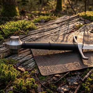 May include: A sword with a polished silver-toned metal pommel and crossguard, featuring a long, cylindrical black grip. The blade is partially visible, extending from the crossguard, with a metallic finish reflecting the surrounding light.
