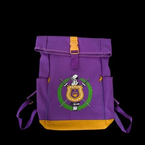 May include: Purple backpack with a yellow accent at the bottom and a yellow buckle. The front of the backpack features a crest with a knight, swords, and a wreath. The backpack has two side pockets with yellow zippers.