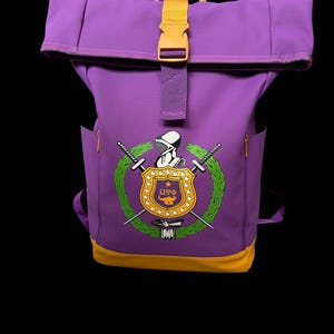 May include: Purple backpack with a yellow buckle and accents. The front features a crest with crossed swords, a wreath, and the Greek letters Omega, Upsilon, and Phi. The bottom of the backpack is yellow.