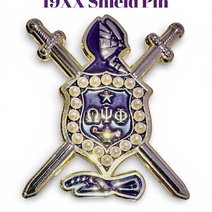 May include: A shield pin with the text "Omega Psi Phi Fraternity 19XX Shield Pin." The pin features a blue and gold design with crossed swords, a knight's helmet, and a star. The pin is surrounded by small, pearl-like accents.