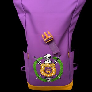 May include: Purple backpack with a yellow accent at the bottom and a yellow buckle. The backpack features a crest with a shield, swords, and a laurel wreath. The crest is in the center of the backpack.