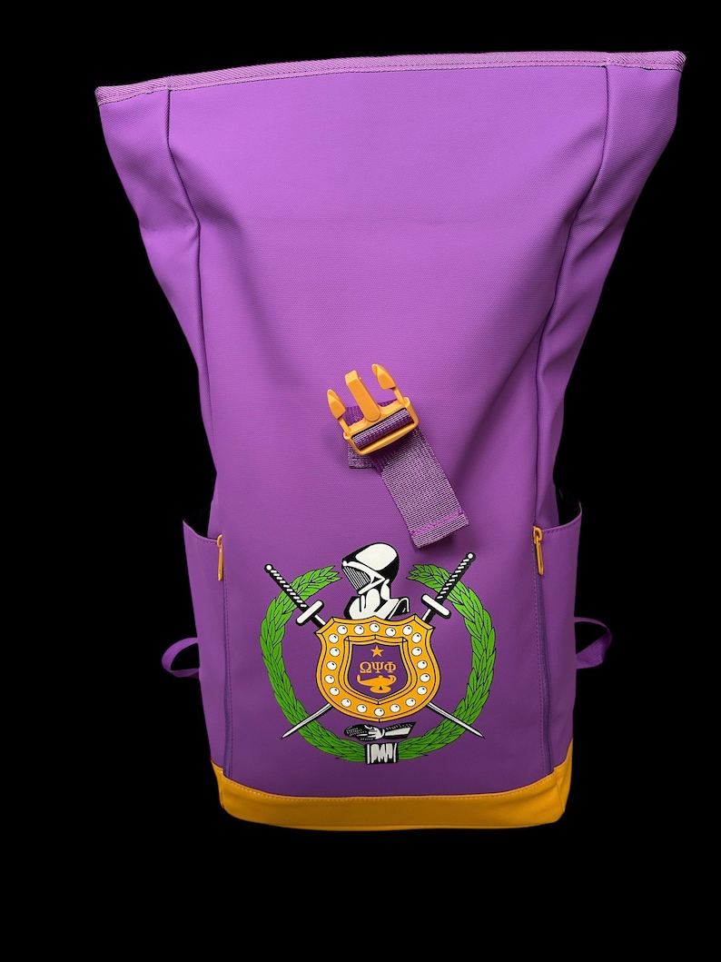 May include: Purple backpack with a yellow accent at the bottom and a yellow buckle. The backpack features a crest with a shield, swords, and a laurel wreath. The crest is in the center of the backpack.