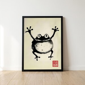 Peut inclure: Une impression d'art encadrée représentant une illustration à l'encre noire d'une grenouille aux bras levés. La grenouille a un corps rond et de grands yeux sur un fond crème. Un petit sceau carré rouge avec des caractères blancs est visible.