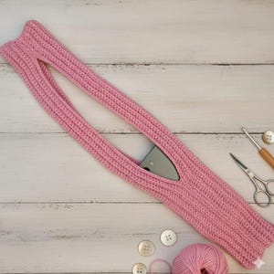 May include: A pink crocheted phone holder with a long strap and a pocket for a phone. The holder is made of a soft, textured yarn. A pair of scissors, a crochet hook, buttons, and a ball of yarn are next to the holder.