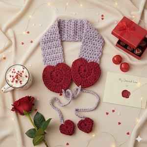 Può includere: Sciarpa all'uncinetto fatta a mano, lavanda e rossa, con due grandi cuori e due piccoli ciondoli a forma di cuore. La sciarpa è esposta su una superficie color crema con decorazioni di San Valentino, tra cui una rosa e una scatola regalo.