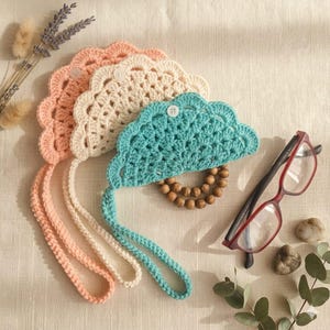 May include: Three semi-circular crochet pouches in peach, cream, and teal, each featuring a single white button closure and a matching crocheted strap. The teal pouch includes a circular wooden bead accent attached to the bottom edge.