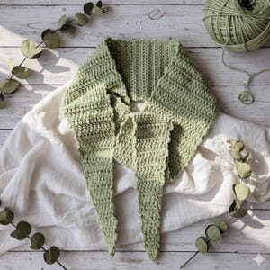 May include: A handmade, light green crochet scarf with a triangular shape and long, pointed ends. The scarf is made of soft yarn and is displayed on a white cloth with eucalyptus branches and a ball of yarn in the background. The scarf is approximately 60 inches long.