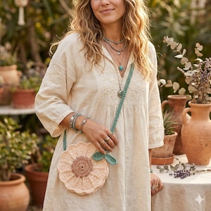 May include: A cream-coloured dress with embroidered details is paired with a crochet sunflower-shaped purse. The purse has a light peach-coloured flower with a brown centre and a green leaf. The purse has a light blue strap.