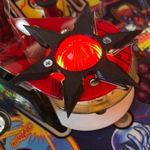 Può includere: Primo piano di un componente del flipper. Un pezzo nero a forma di stella circonda una luce rossa brillante. Lo sfondo presenta grafiche colorate e la parola "KA-WING".