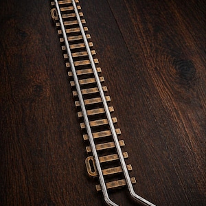 Könnte beinhalten: Ein Abschnitt einer Modelleisenbahnstrecke mit Holzschwellen und silbernen Schienen. Die Strecke hat ein metallisches, gealtertes Aussehen. Auf dunklem Holzmaserungshintergrund.