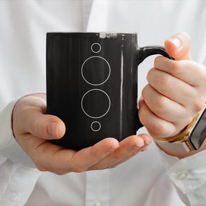 Puede incluir: Una taza de cerámica negra con una disposición vertical de cuatro contornos circulares blancos de diferentes tamaños centrados en la parte frontal. La taza tiene un acabado liso y brillante y un asa curva en el lado derecho para sujetarla.