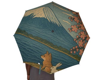 May include: An open umbrella with a Japanese-inspired design. The umbrella features a mountain, lake, and cherry blossom illustration. A brown dog and white rabbit are sitting on the shore. The umbrella is held by a person wearing a gray shirt.