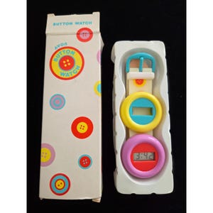 Può includere: Un orologio Avon Button Watch vintage nella sua confezione originale. L'orologio ha un cinturino bianco e tre componenti colorati a forma di bottone: giallo, blu e rosa. Il quadrante mostra l'ora. La scatola è bianca con grafica a bottoni e la scritta "BUTTON WATCH".