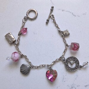 May include: A silver-toned charm bracelet featuring a toggle clasp and various charms including a clock face, a heart-shaped pink crystal, a seashell, a rectangular floral metal piece, a square bead, and several translucent pink glass beads.