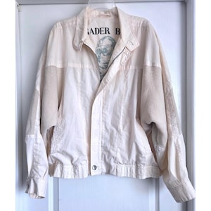 May include: A cream-colored zip-up jacket featuring a collar, long sleeves with ribbed panels, and a front zipper closure. The interior lining displays the text "RADER B" above a graphic illustration of a person wearing goggles and a helmet.