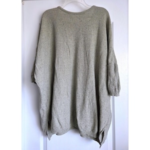 May include: A light green, oversized sweater with a round neckline and three-quarter sleeves. The sweater has a textured knit and a relaxed fit. The garment is hanging on a white hanger against a white door.