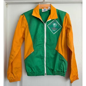 May include: A vintage green and orange zip-up jacket with a white zipper. The jacket features a diamond-shaped patch with the text "OLYMPIC 1984". The sleeves and side panels are orange, while the front and back are green.
