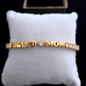 May include: A gold-colored bracelet with the word "GRAND MOM" spelled out in individual letters. The bracelet has a small, clear gem in the center and additional gems along the band. The bracelet is displayed on a white, square pillow.