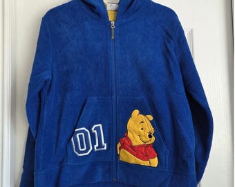 May include: A blue zip-up hoodie featuring a Winnie the Pooh graphic on the front. The hoodie has a front pocket with the number "01" and a hood. The zipper is silver. The interior of the hood is yellow.