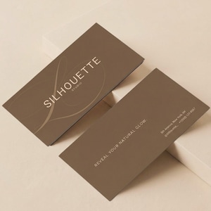 Puede incluir: Dos tarjetas de visita rectangulares de color marrón oscuro con texto en beige claro. Una tarjeta muestra el texto SILHOUETTE STUDIO con un gráfico circular, mientras que la otra presenta el texto REVEAL YOUR NATURAL GLOW e información de contacto.
