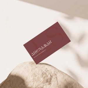 Puede incluir: Una tarjeta de visita rectangular de color granate oscuro con texto en blanco que dice ARENAMAR y Exclusive handmade art. La tarjeta está colocada en ángulo, apoyada contra un objeto texturizado, de color beige claro y forma redondeada.