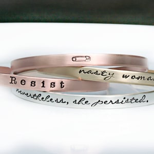 May include: Three stacked metal bracelets with the words "Resist", "nasty woman", and "nevertheless, she persisted." The top bracelet is rose gold, the middle bracelet is silver, and the bottom bracelet is silver.