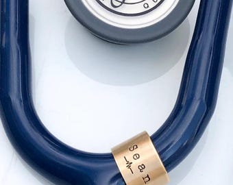以下が含まれることがあります： ネイビーブルーの聴診器で、チューブにメタリックゴールドの識別リングが取り付けられています。ゴールドのリングには「Sean」という名前と小さな心拍のパルスラインのグラフィックが刻印されています。聴診器のチェストピースが上部に見えています。