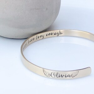 May include: Gold bangle bracelet with the inscription "I have been long enough" and the name "Olivia" with angel wings.