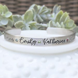 May include: Silver bangle bracelet with the names "Aliban", "Emily", "Katherine", and "Erin" engraved on it. The bracelet has four small gemstones set in the metal.