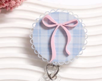 May include: A circular badge reel featuring a light blue and white plaid pattern background with a scalloped edge. A soft pink ribbon bow is centred on the front of the badge. A clear plastic strap with a metal snap button is attached to the bottom.