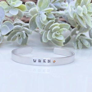 May include: A silver-toned metal cuff bracelet with an open back design. The front of the bracelet is stamped with the name WREN in black capital letters, followed by a small, circular, amber-colored gemstone accent set into the metal surface.