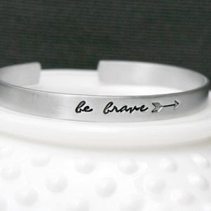 May include: A silver-toned metal cuff bracelet with a brushed finish. The exterior surface features the engraved text "be brave" in a cursive script, followed by a small arrow symbol pointing to the right. The bracelet has an open-ended design.
