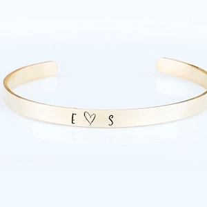 May include: A gold bangle bracelet with the letters "E" and "S" engraved on it, separated by a heart symbol.