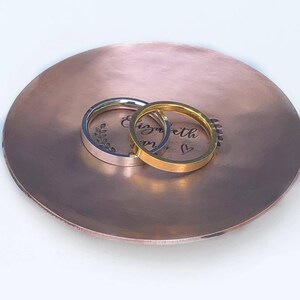 May include: A round, hammered copper-toned dish featuring the engraved name Elizabeth and a small heart symbol. Two smooth, flat-band rings, one silver-toned and one gold-toned, are resting on the surface of the dish.