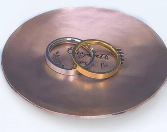 May include: A round, hammered copper-toned dish featuring the engraved name Elizabeth and a small heart symbol. Two smooth, flat-band rings, one silver-toned and one gold-toned, are resting on the surface of the dish.