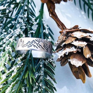 May include: A silver-toned metal ring featuring a laser-engraved design of a mountain range, a rising sun, and evergreen trees. The band has a smooth, polished finish and a flat profile, showcasing a detailed landscape scene around the exterior.