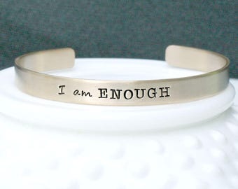 May include: A silver-toned metal cuff bracelet with a brushed finish. The front of the bracelet is stamped with the black text "I am ENOUGH" in a combination of script and block lettering. The bracelet features an open back design.