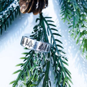 May include: A silver-toned metal ring featuring a continuous engraved pattern of small evergreen trees around the outer band. The surface of the ring has a brushed finish, and the tree designs are darkened to provide contrast against the metal.