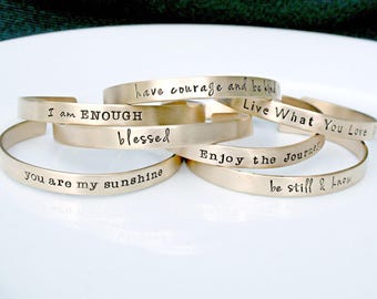 May include: A collection of gold-toned metal cuff bracelets, each featuring stamped black text. The phrases include have courage and be kind, Live What You Love, I am ENOUGH, blessed, Enjoy the Journey, you are my sunshine, and be still & know.
