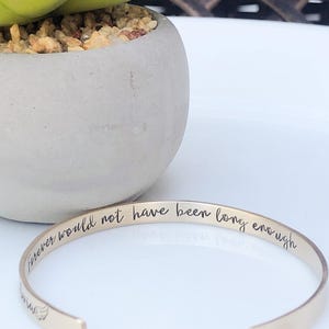 May include: A gold-toned metal cuff bracelet featuring an open design. The interior surface is engraved with the black cursive text "Forever would not have been long enough" and includes a small graphic of a wing on the left side of the band.
