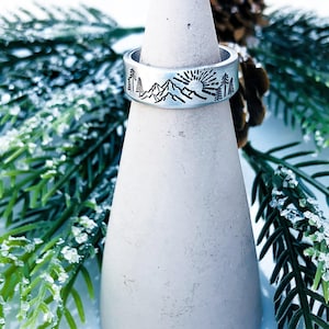 May include: A silver-toned metal ring featuring a stamped black graphic design that depicts a mountain range, a rising sun, and several pine trees. The band has a smooth, flat surface and is displayed on a conical stand for clear visibility.