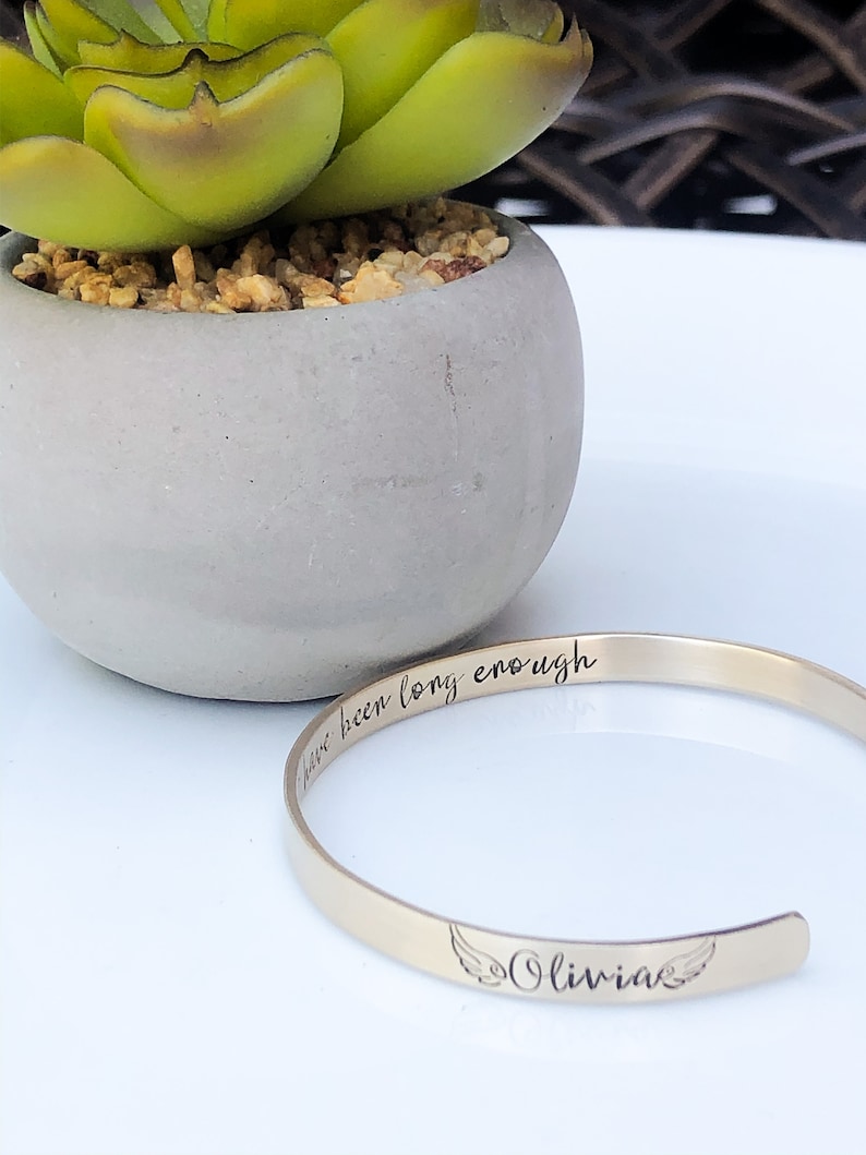 May include: Gold bangle bracelet with the inscription "I have been long enough" and the name "Olivia" with angel wings.