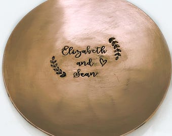 May include: A round, metallic copper-colored dish featuring a brushed finish. The center is engraved with the text "Elizabeth and Sean" in a cursive script, flanked by two small, delicate leaf branch designs and a heart symbol.