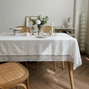 May include: A white rectangular tablecloth with a decorative lace border, set on a wooden table. The table is set with a vase of white flowers, candles, and a cake stand. The room has a neutral color palette.