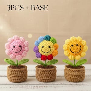 Könnte beinhalten: Drei gehäkelte Blumendekorationen mit lächelnden Gesichtern. Eine ist rosa, eine ist regenbogenfarben und eine ist gelb. Jede Blume sitzt in einem braunen, gehäkelten Topf mit grünen Blättern und einem braunen Stiel. Der Text "3PCS + BASE" steht oben.